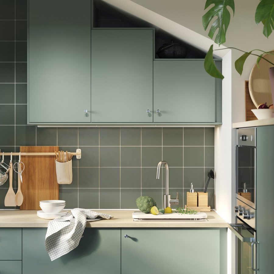Grey-green kitchen cabinets stepped under a sloping ceiling, with a tiled wall and a pale worktop